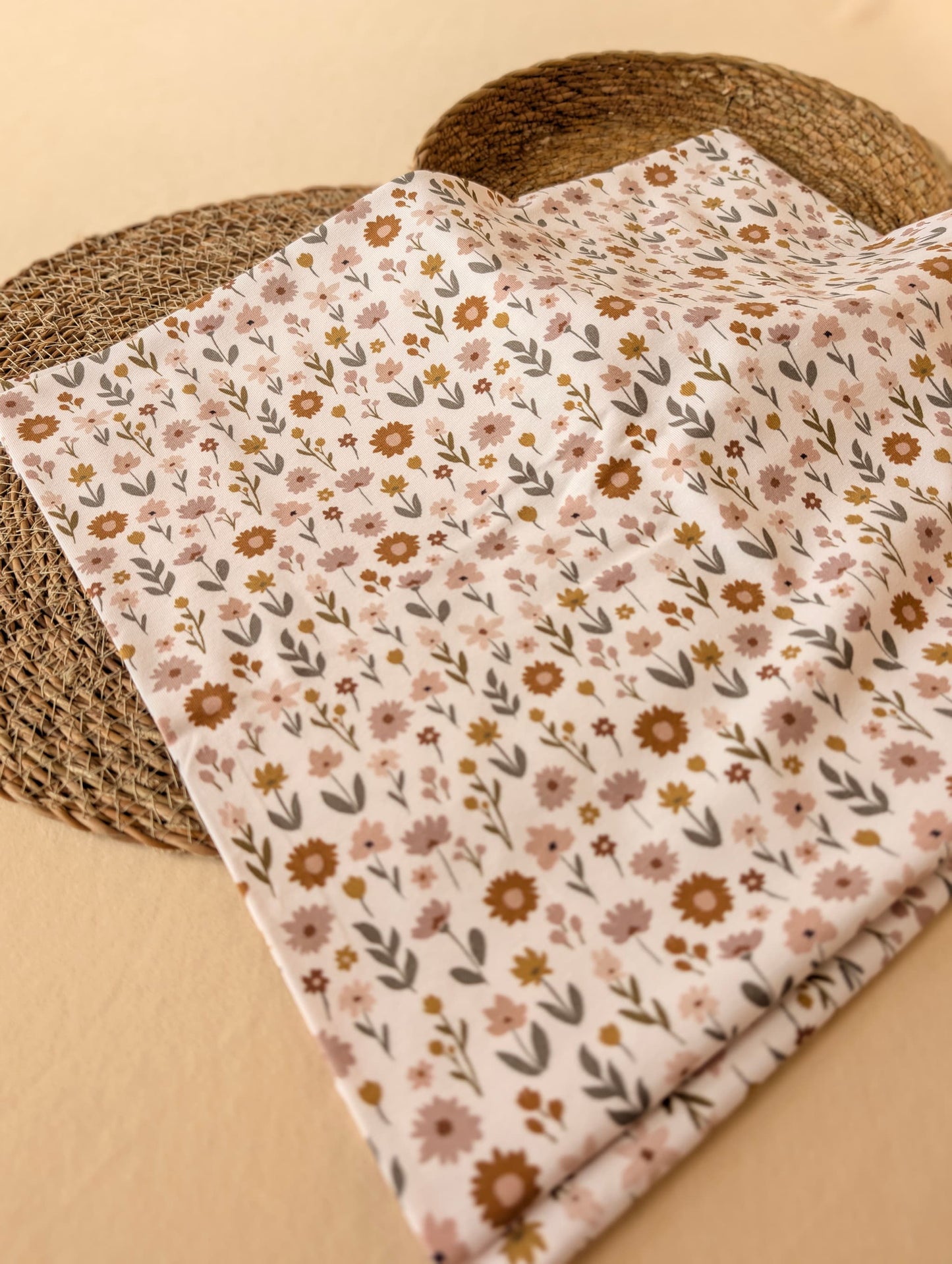 Ottie Floral cotton jersey folded flat on wicker showing the scattered wildflower print — daisies and botanical sprigs in dusty pink, mustard and sage on ivory — Esorai