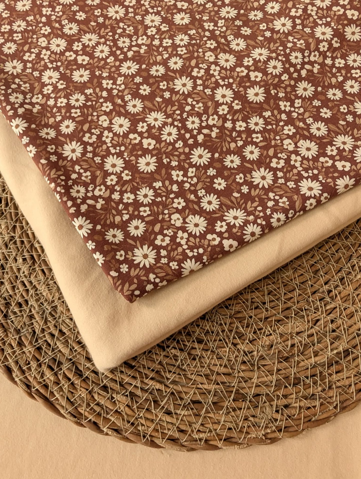 Willa Floral Cotton Jersey in Rust — folded fabric showing dense white daisy print on warm brown jersey with natural cream reverse, on woven rattan