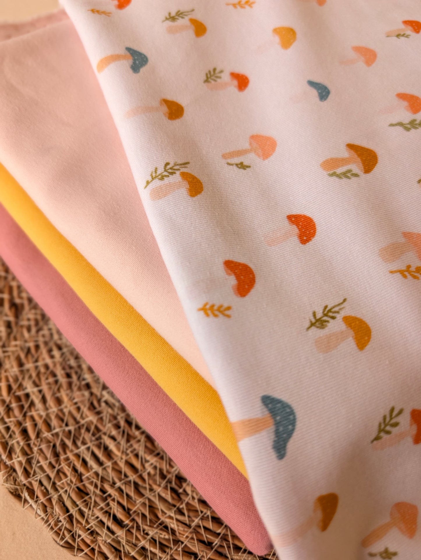 Cotton rib fabric draped on rattan showing scattered toadstool and botanical sprig print — stacked with plain colours to match