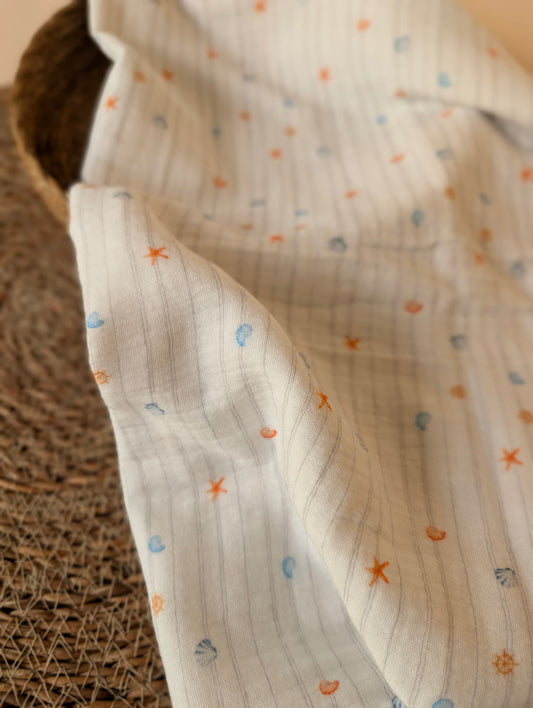 White double gauze fabric draped over a wicker basket, Cora Coastal print featuring scattered starfish, scallop shells and ship's wheel motifs in orange and blue — Esorai