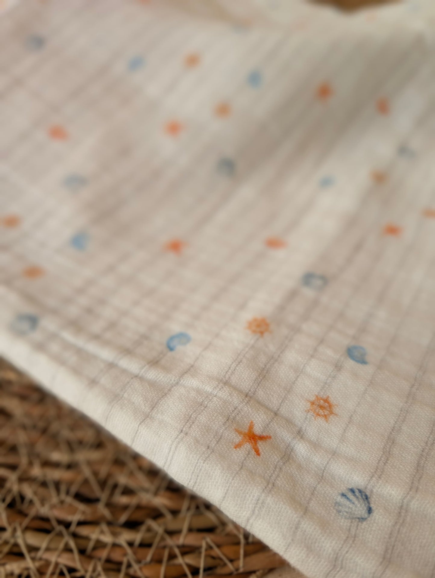 Double gauze fabric folded on a wicker mat showing Cora Coastal print — tiny starfish, shells and nautical motifs in coral and blue on white — Esorai