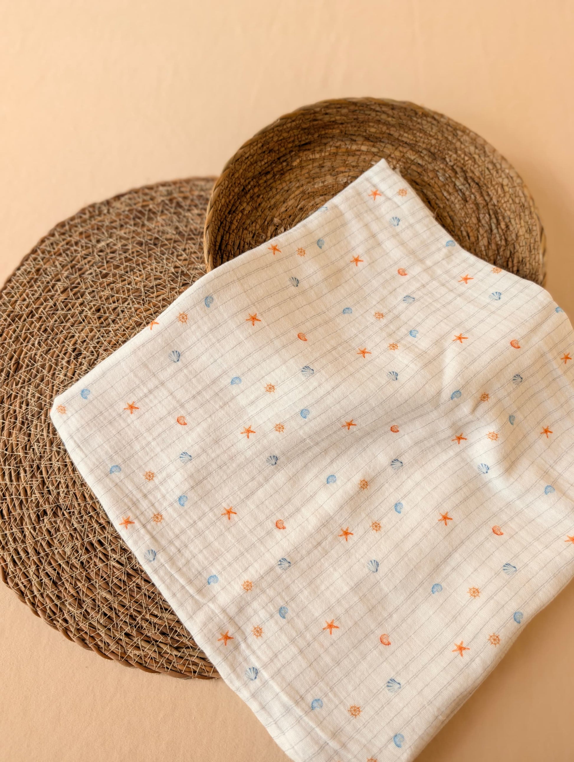 Double gauze fabric folded on a wicker mat showing Cora Coastal print — tiny starfish, shells and nautical motifs in coral and blue on white — Esorai