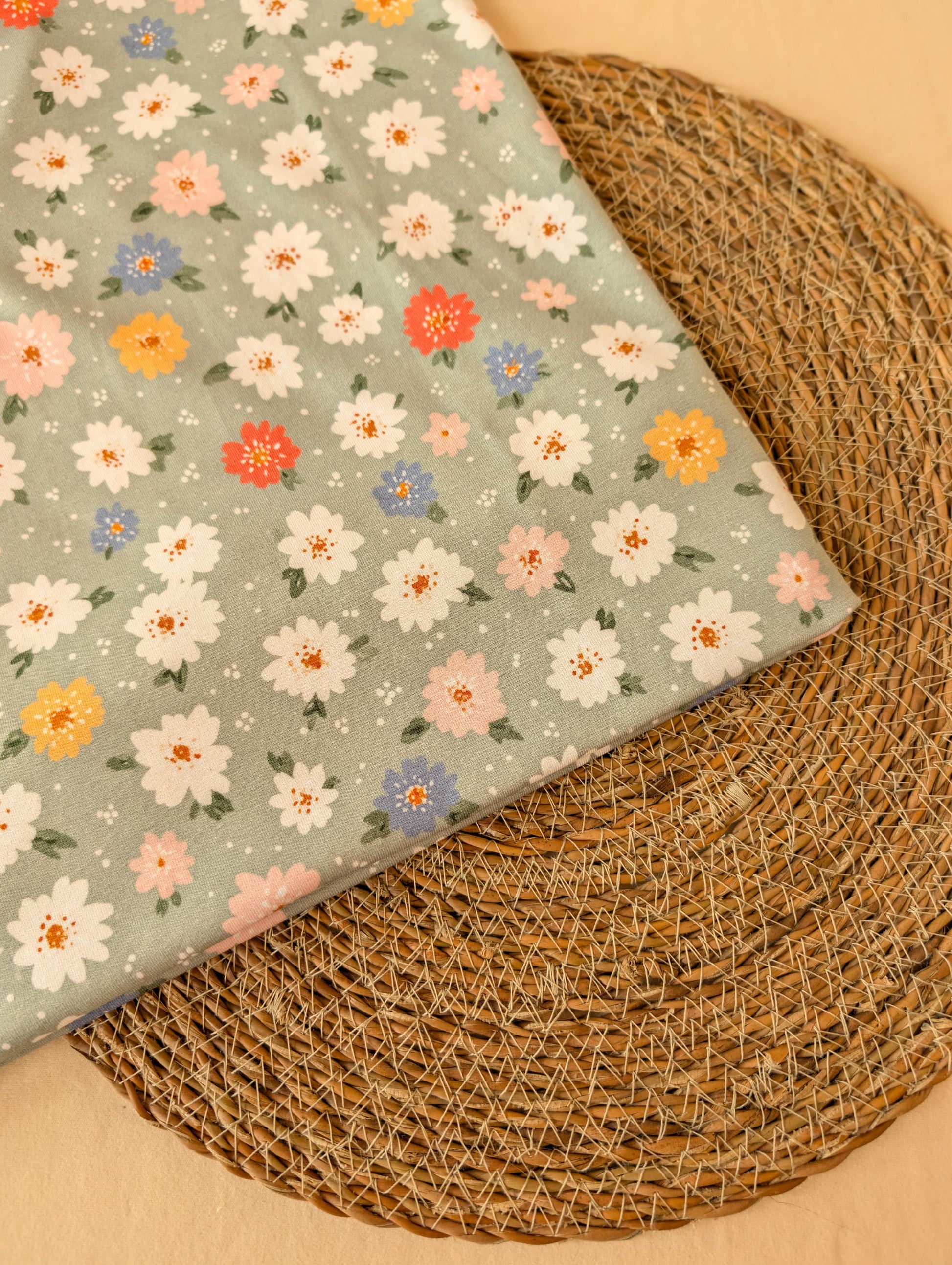 
Daisy Floral Cotton Jersey Sage folded on wicker mat — angled view of daisy print on soft sage green jersey ground — Esorai