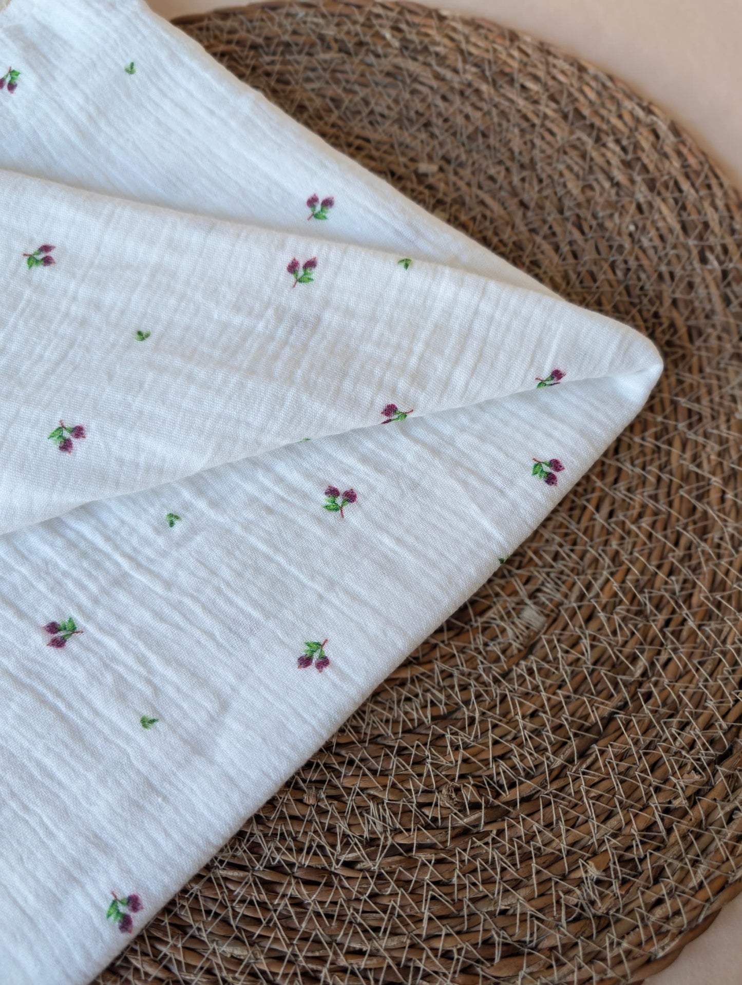 Double gauze fabric folded on a rattan placemat showing the full Mini Blueberries print layout — tiny plum berry clusters on white