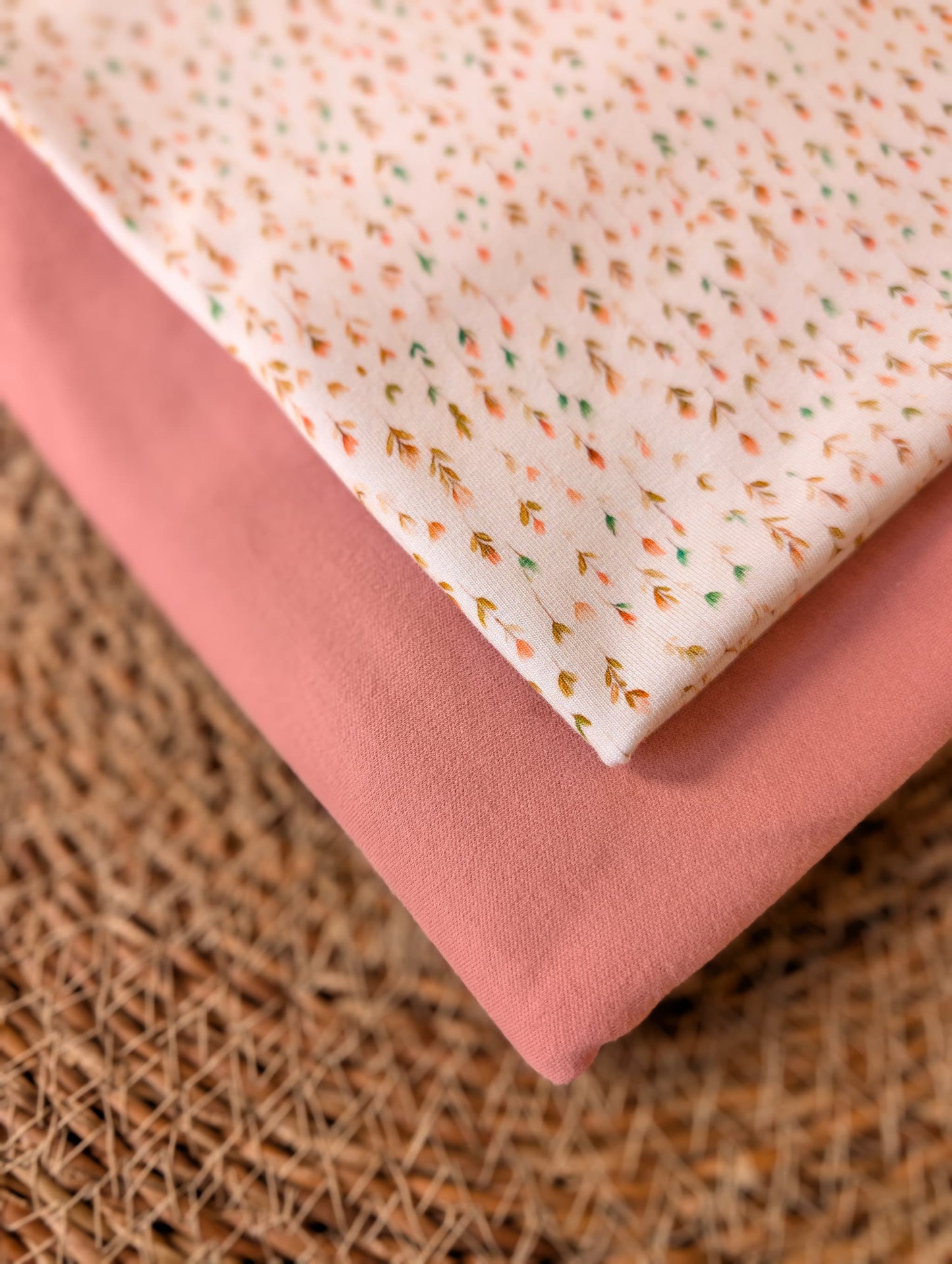 Cotton jersey Edie Sprig fabric stacked with plain dusty pink cotton jersey on a rattan placemat — fine scattered sprig print in peach, mustard and sage on white