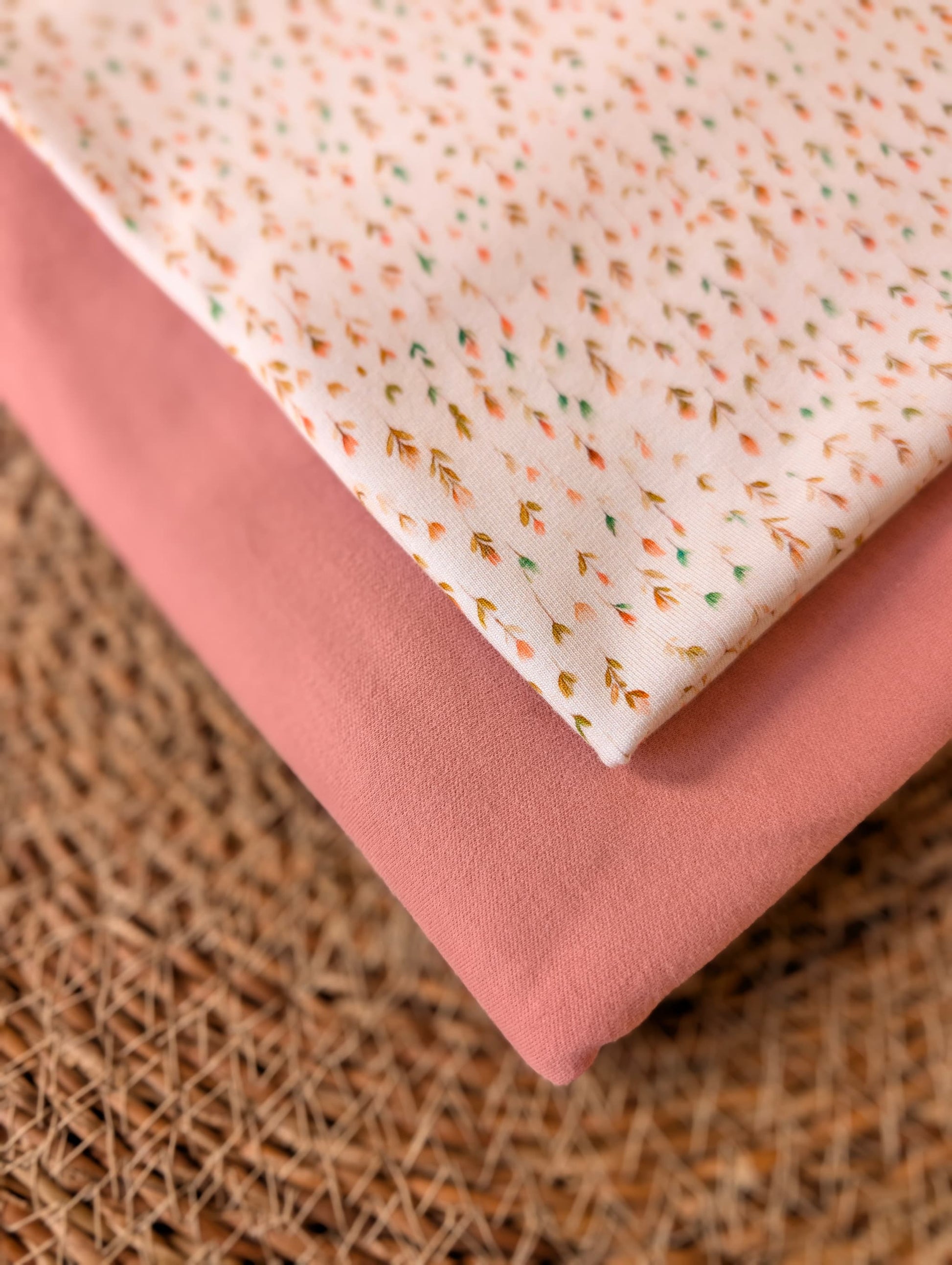 Cotton jersey Edie Sprig fabric stacked with plain dusty pink cotton jersey on a rattan placemat — fine scattered sprig print in peach, mustard and sage on white