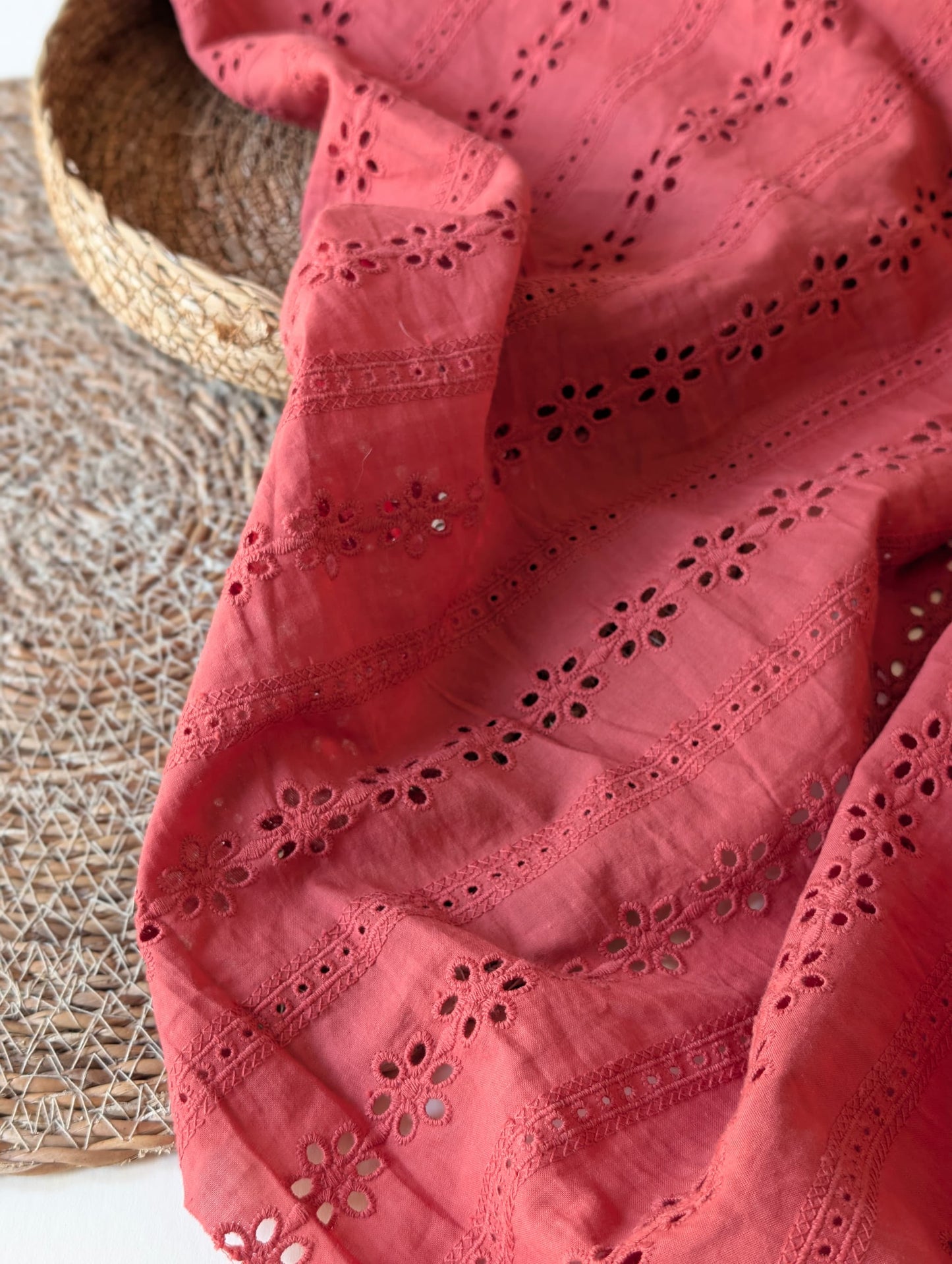 Rust/terracotta cotton broderie anglaise fabric draped over a woven basket, showing rows of floral eyelet embroidery with ladder-stitch trellis borders