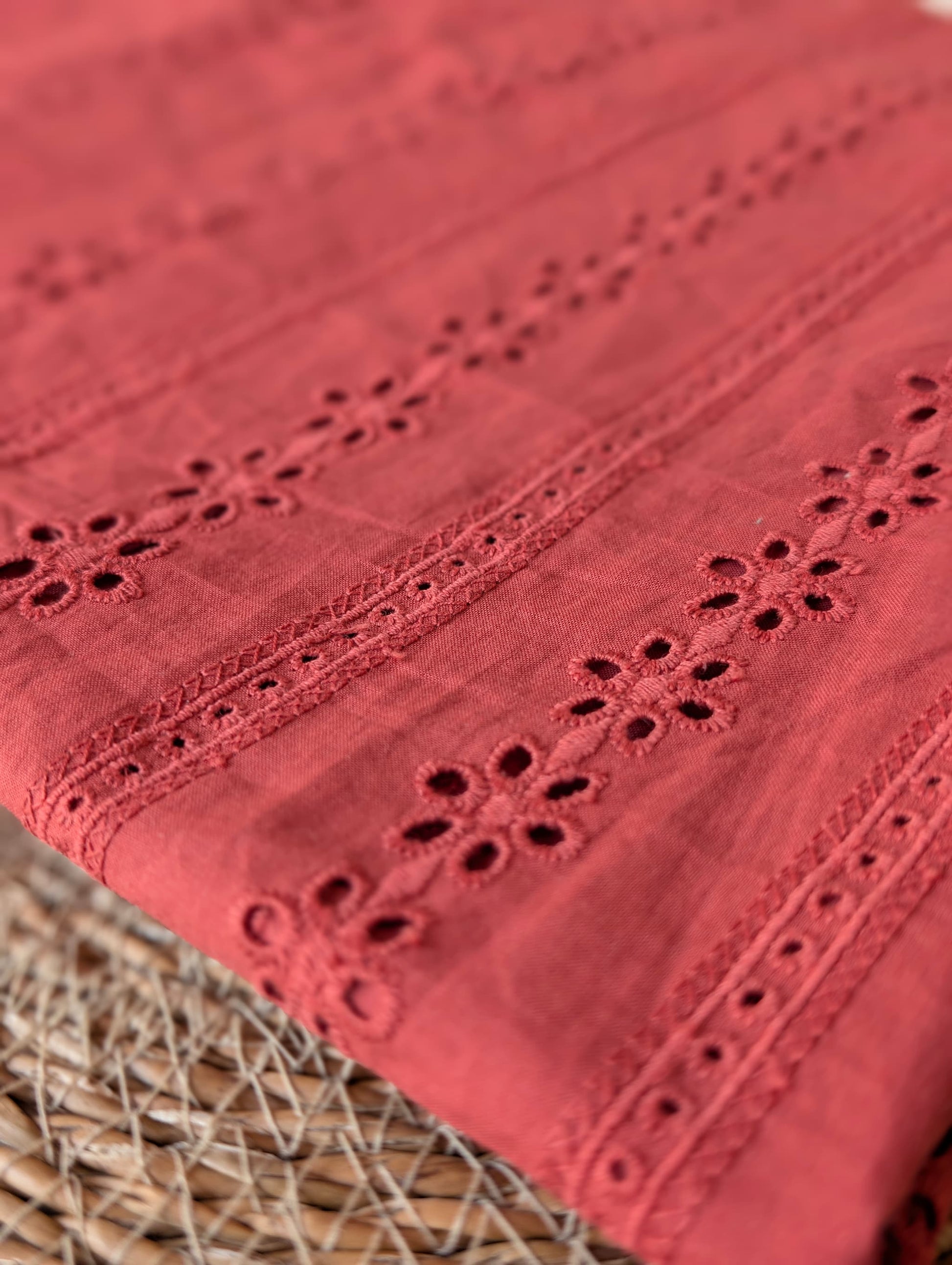 Close-up of Esme cotton broderie anglaise fabric folded diagonally, showing the rust/terracotta floral eyelet embroidery and ladder-stitch trellis detail