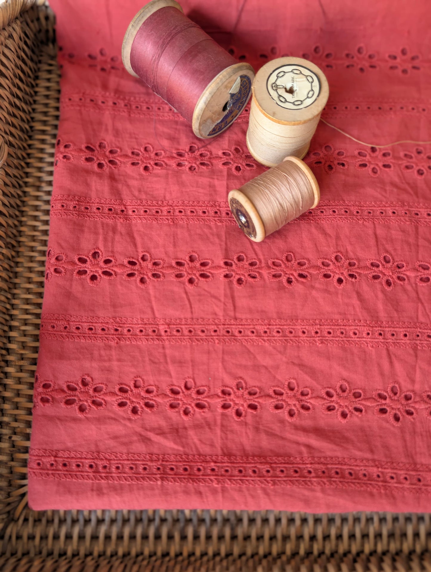 Esme cotton broderie anglaise fabric folded flat with vintage wooden thread spools resting on top, showing the rust/terracotta floral eyelet embroidery stripes