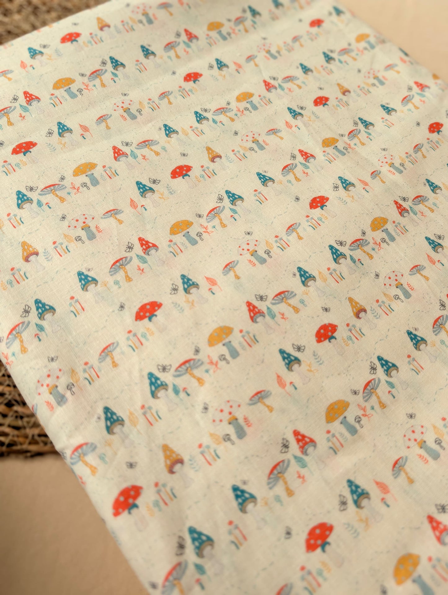Linen cotton fabric folded flat showing the full Hazel Woodland print — detailed toadstools, butterflies and botanicals in red, teal, mustard and grey on natural