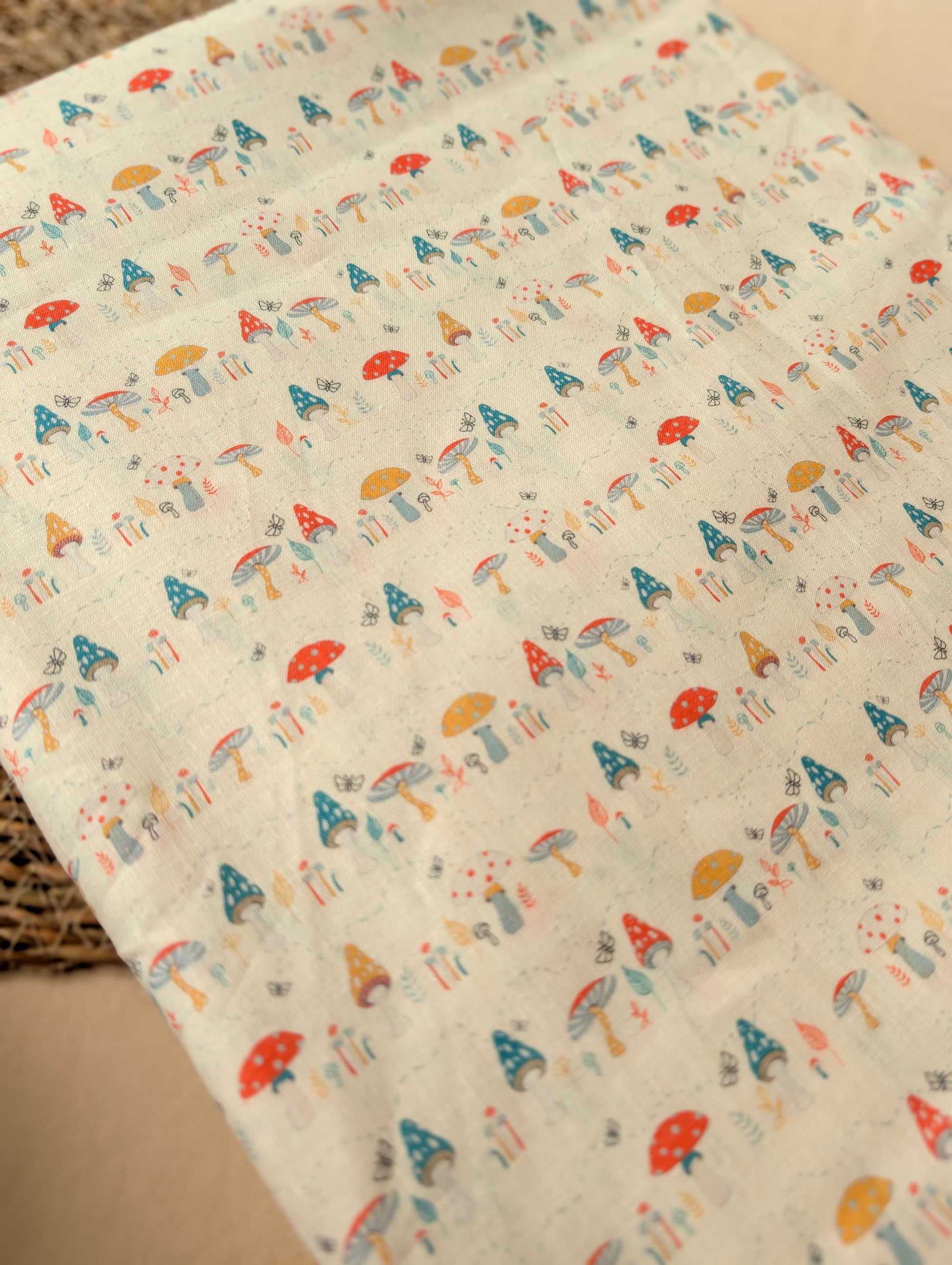 Linen cotton fabric folded flat showing the full Hazel Woodland print — detailed toadstools, butterflies and botanicals in red, teal, mustard and grey on natural