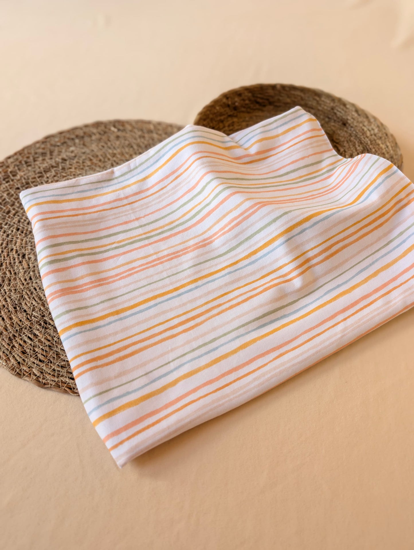 Cotton jersey fabric folded on a rattan placemat — Isla Stripe in pastel multi, with hand-drawn stripes in peach, mustard, sage and duck egg on white