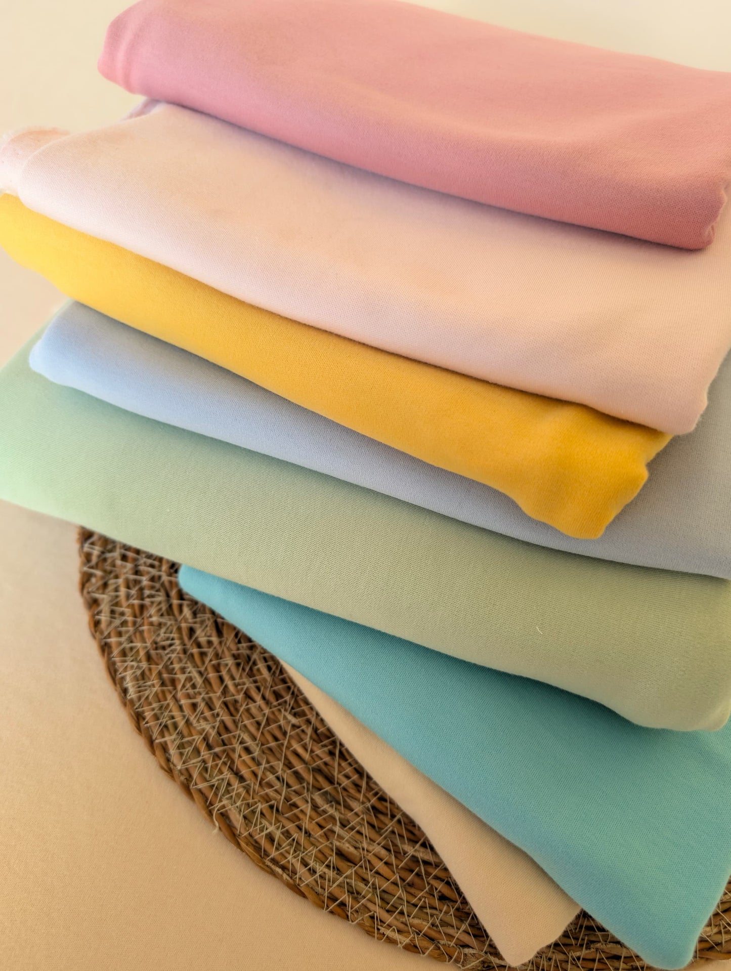 Stack of six premium-weight organic cotton jersey fabrics in Dusky Rose, Blush, Buttercup, Powder, Pistachio and Lagoon, folded on a wicker mat — Plain Cotton Jersey collection by Esorai
