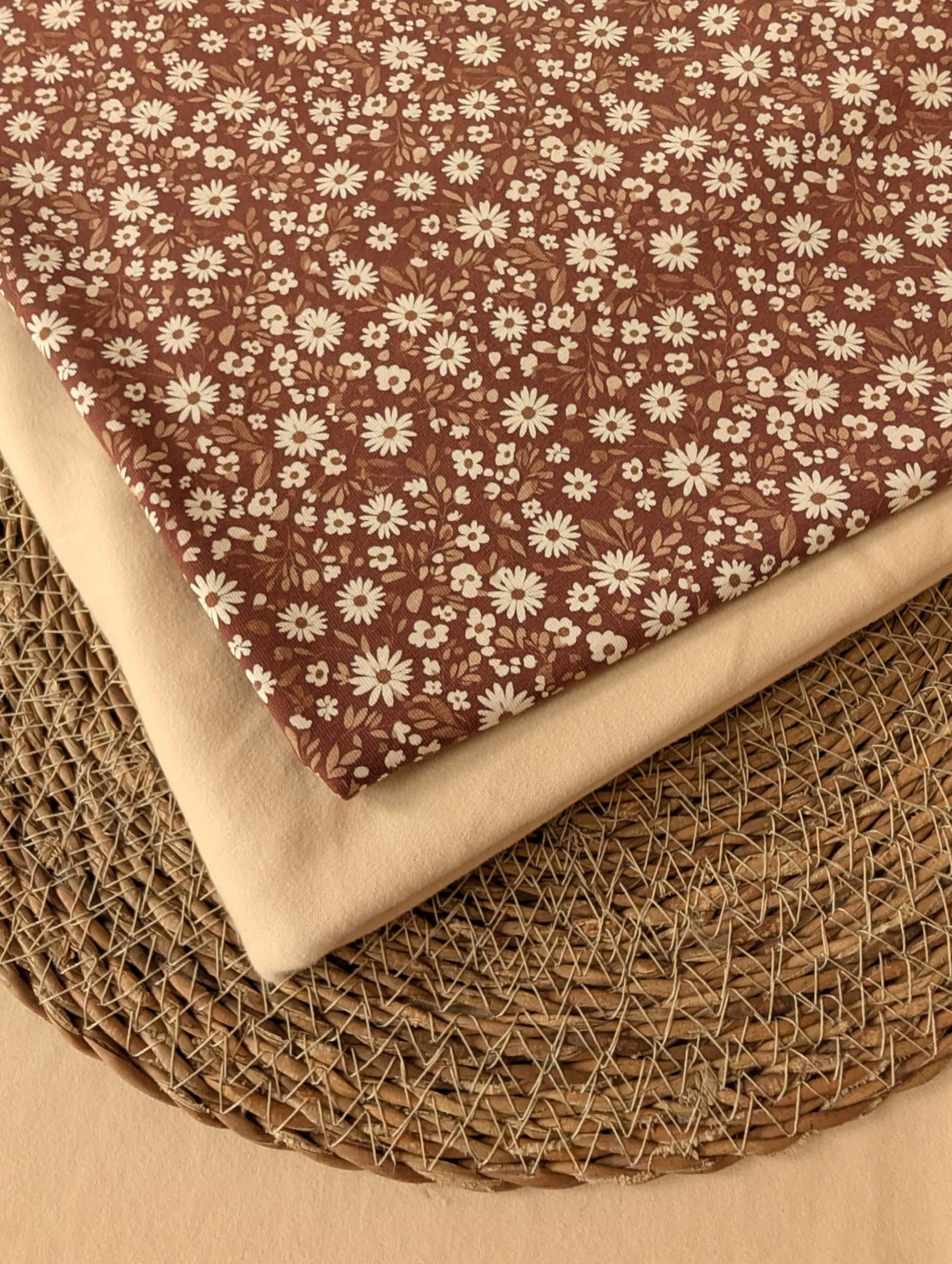 Willa Floral Cotton Jersey in Rust — folded fabric showing dense white daisy print on warm brown jersey with natural cream reverse, on woven rattan