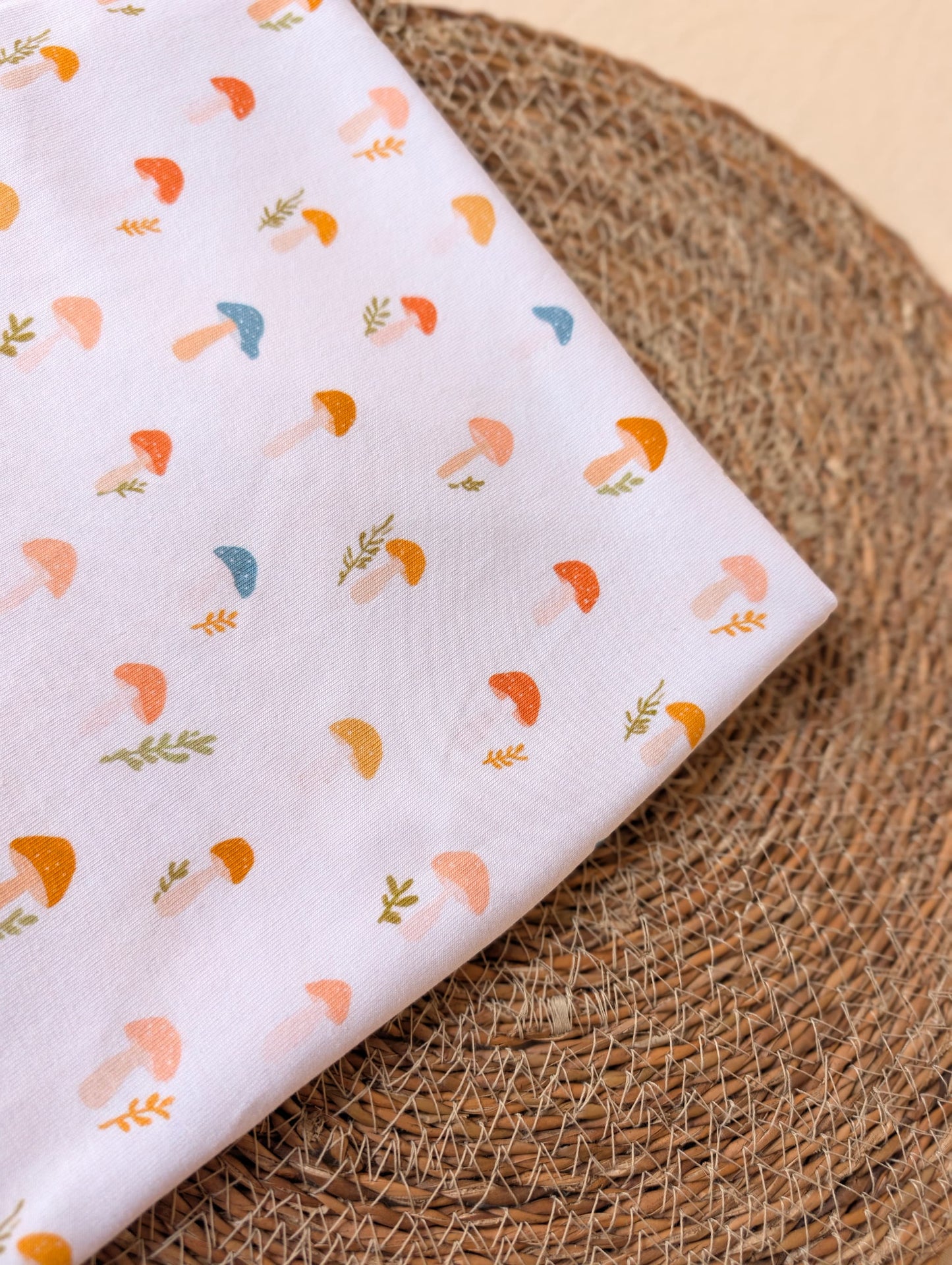 Cotton rib fabric folded on a rattan placemat showing full layout of Wren toadstool print — scattered mushrooms and sprigs in terracotta, mustard, teal and peach on white